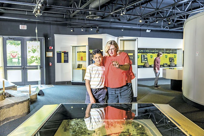 mom and sun looking at an informative exhibit inside the Fort Ancient Museum with dad standing and mom kneeling next to a toddler inside the Native American structure at Fort Ancient Earthworks & Nature Preserve in Oregonia Ohio